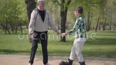 可爱的男孩穿着格子衬衫在公园里滑旱冰，他的祖父抓住他拥抱。 户外活动